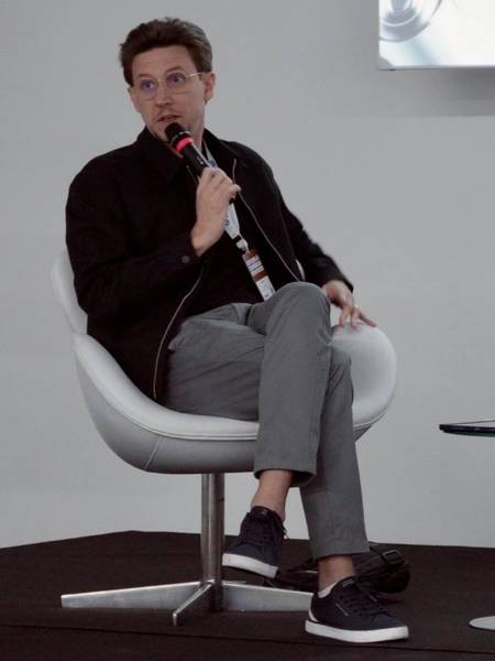 Léo Andrade sentado no palco, falando ao microfone durante um painel