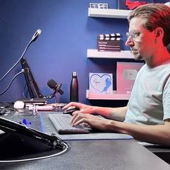 Léo Andrade trabalhando no estúdio, digitando, com o microfone e a placa do YouTube ao fundo