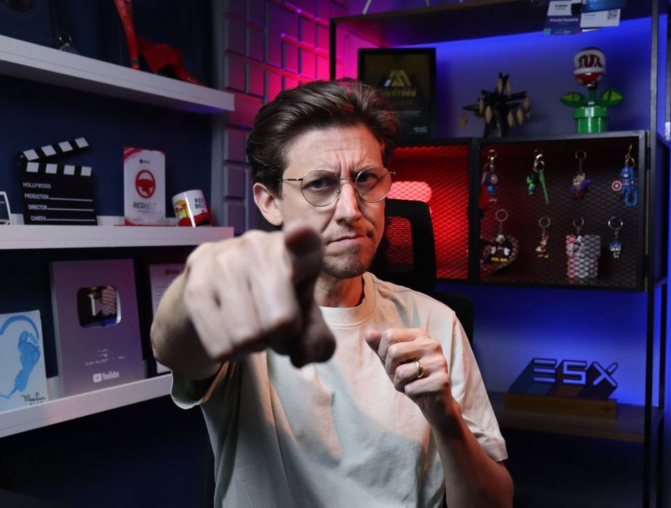 Léo Andrade gravando no estúdio, apontando para a câmera, com a placa de prata do YouTube e a estante iluminada ao fundo